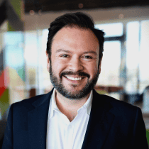 Man with dark hair and beard in white shirt, dark blazer, smiles in office styled like Chuck E. Cheese arcade and kids birthday party.