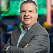 Middle-aged man in grey suit stands smiling with arms crossed at Chuck E. Cheese, colorful arcade games and lights behind him.
