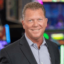 A man in a suit jacket and light shirt smiles at Chuck E. Cheese, with arcade games and kids’ birthday party lights in the background.