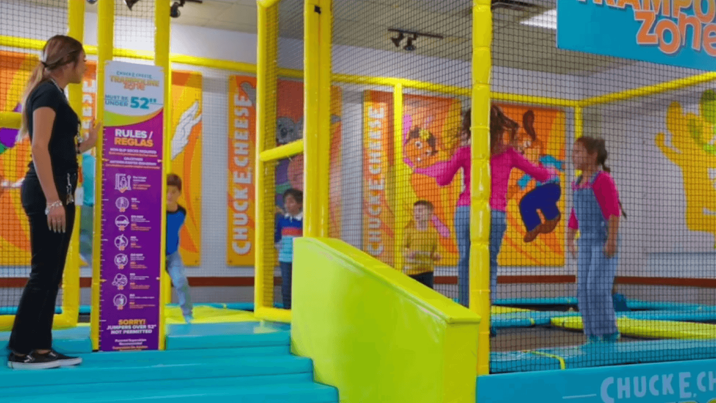 Kids jump in an enclosed trampoline area at Chuck E. Cheese near arcade games, wall art, and birthday party decorations.