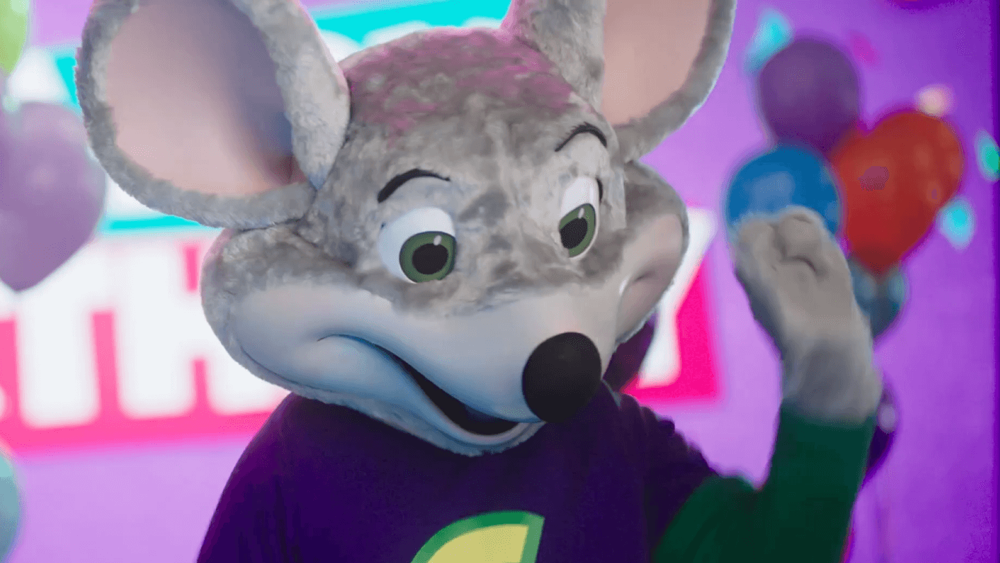 Chuck E. Cheese mascot in purple shirt waves by balloons, family fun center, birthday party for kids, arcade games, pizza and Fun Pass.