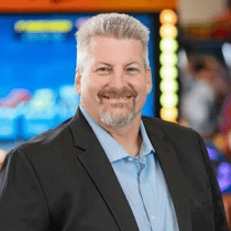 Smiling man with gray hair stands by kids' arcade games at Chuck E. Cheese family fun center during a birthday party.