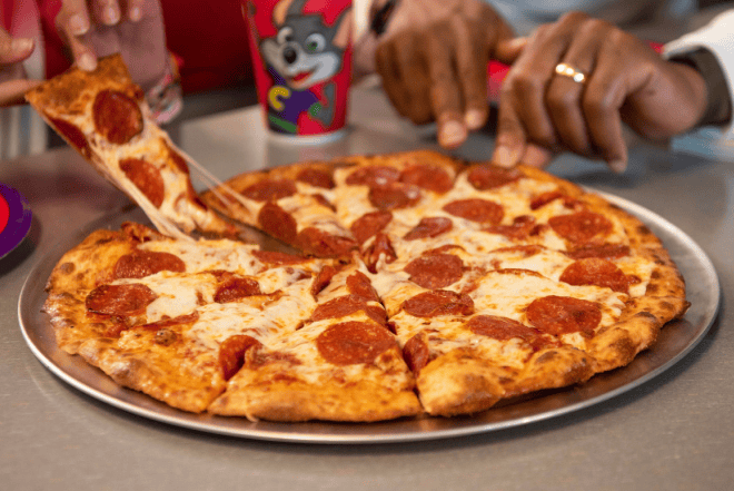 Kids at Chuck E. Cheese share pepperoni pizza; a Chuck E. cup in background, hands visible during birthday party with arcade games.