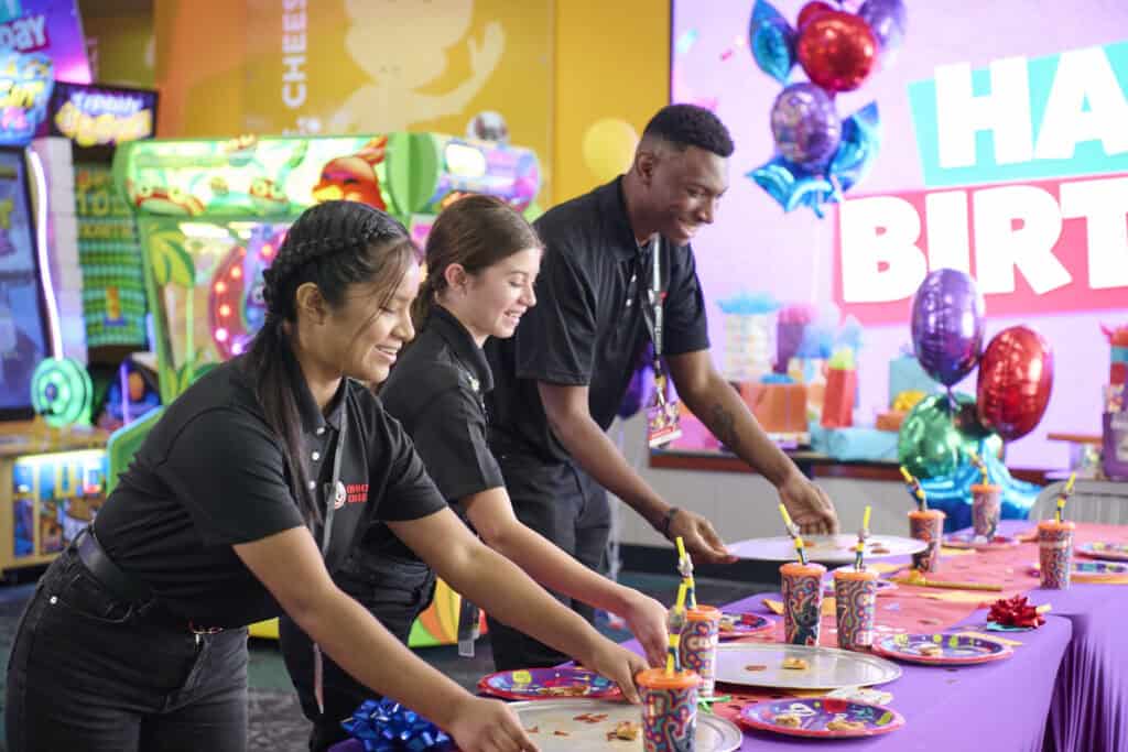 Employees set up a kids birthday party table with plates and decorations at Chuck E. Cheese, arcade games and balloons nearby.