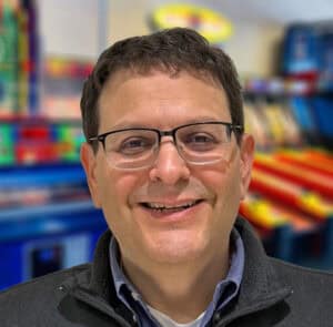 Man with glasses and short brown hair smiles in front of Chuck E. Cheese arcade games, Fun Pass, and kids entertainment area.