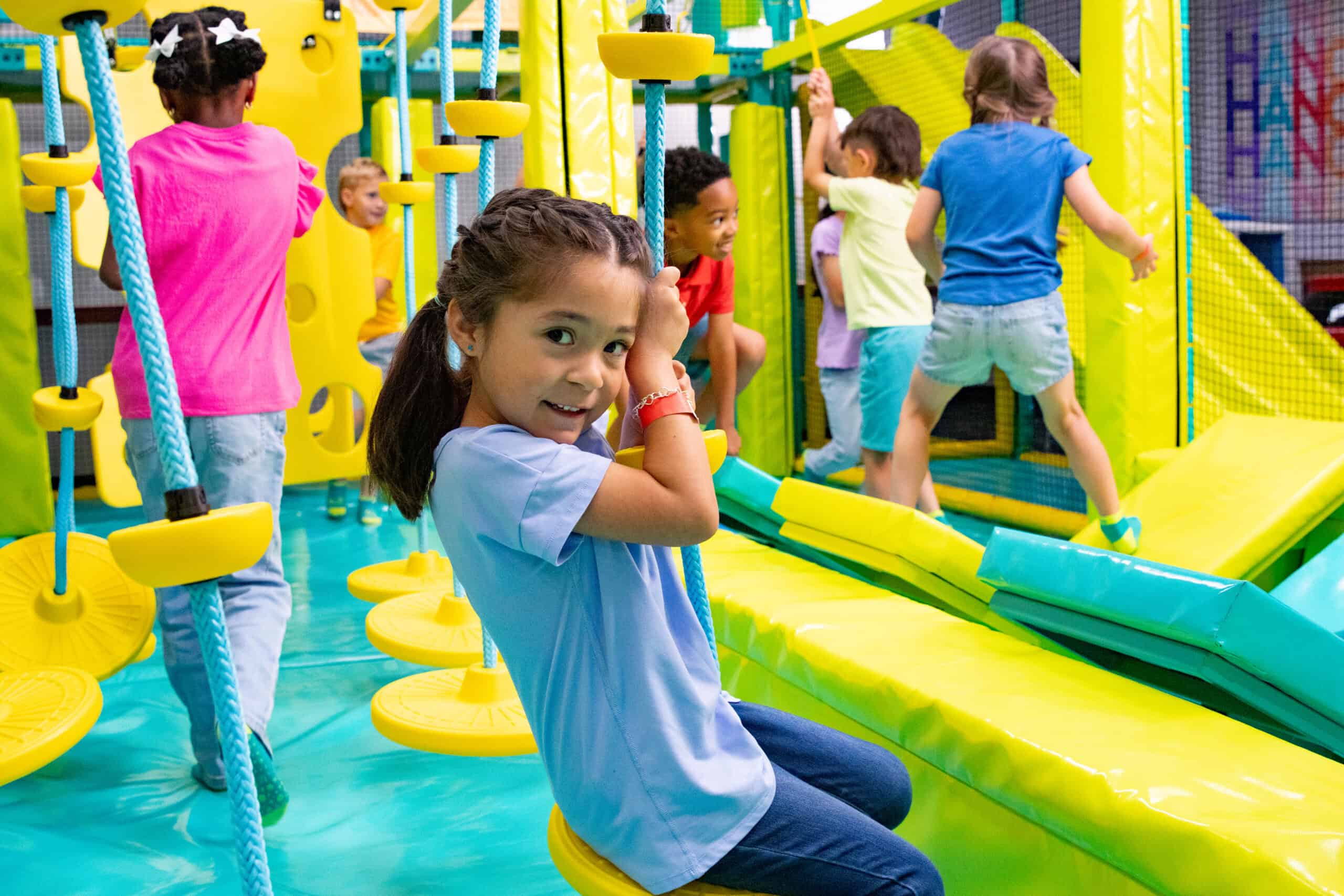 A young girl on a rope swing at Chuck E. Cheese, with kids climbing colorful play equipment in the background.