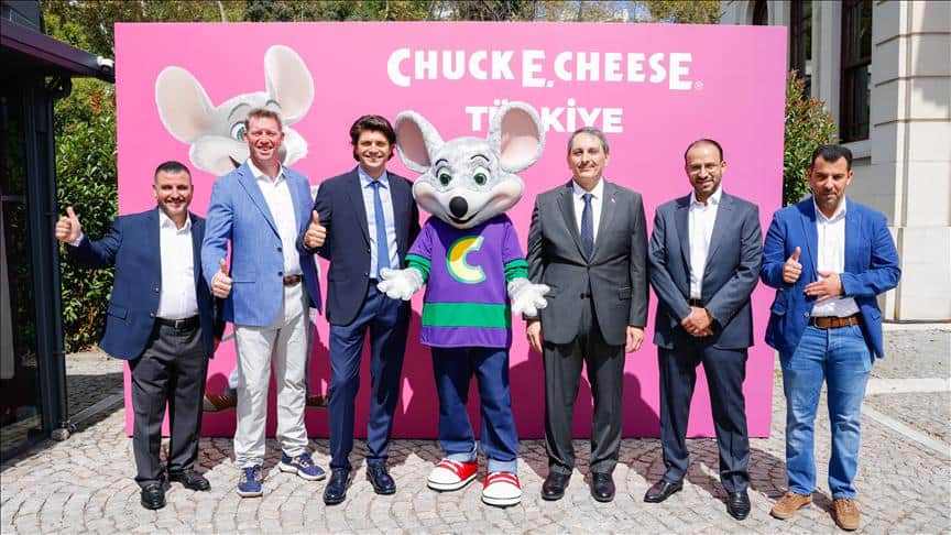Six men in business attire with Chuck E. Cheese mascot at Türkiye family fun center backdrop, thumbs up for kids birthday party and games.