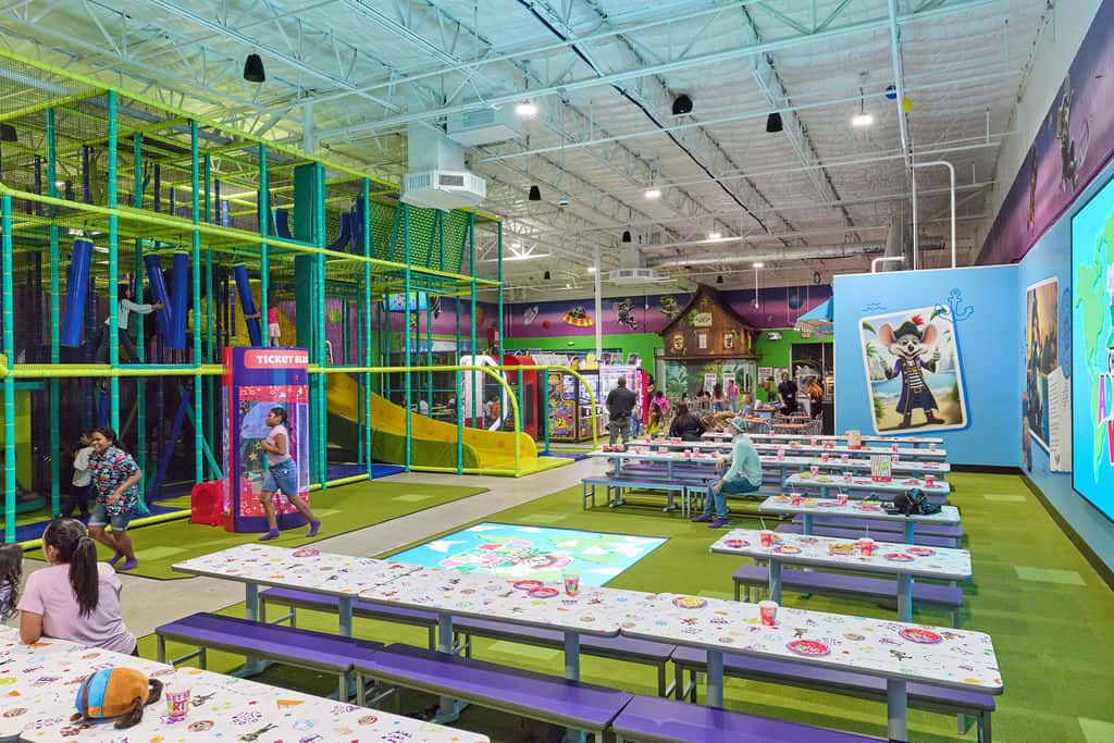 Chuck E. Cheese fun center showing climbing structures, slides, arcade games, birthday party tables, and kids playing.