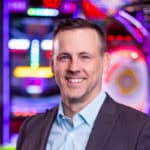 A man in a suit jacket smiles in front of arcade games for kids at Chuck E. Cheese family fun center, known for pizza and birthday parties.