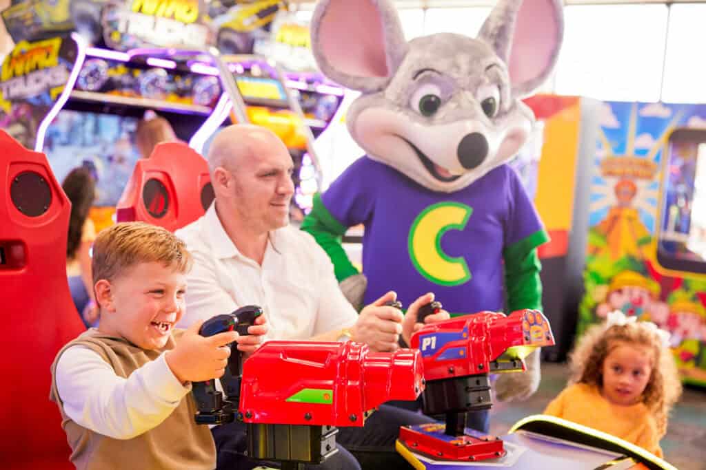 Chuck E. Cheese mascot stands behind family at arcade shooting game, with kids winning tickets