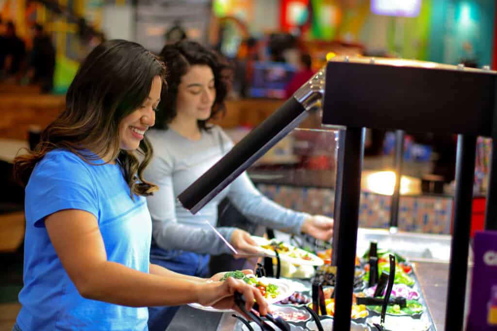 Two women choose fresh vegetables at the Chuck E. Cheese salad bar during a kids birthday party near arcade games and pizza.