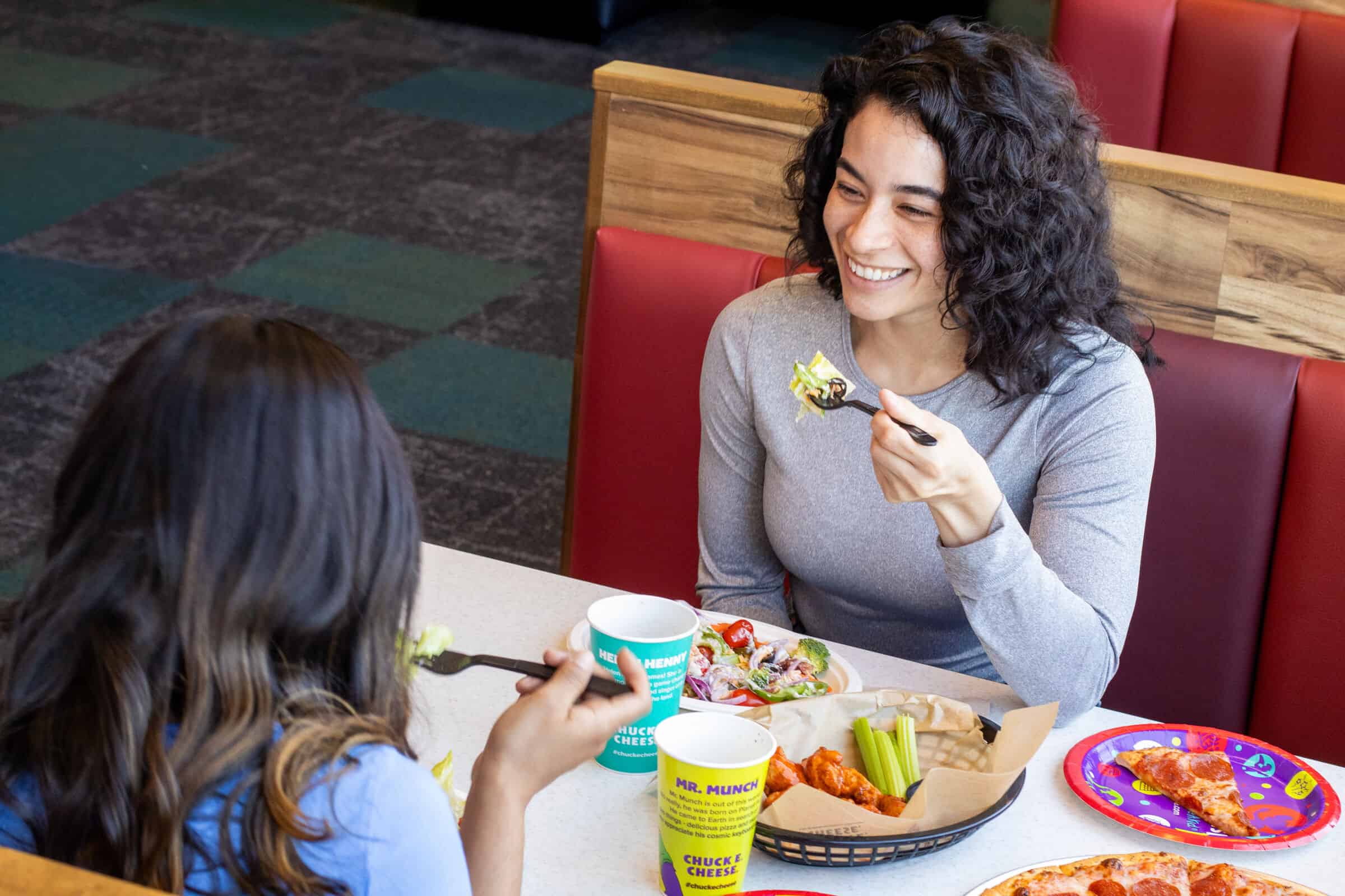 Two women at a Chuck E. Cheese booth share pizza, salad, and wings during a kids birthday party; arcade games in the background.