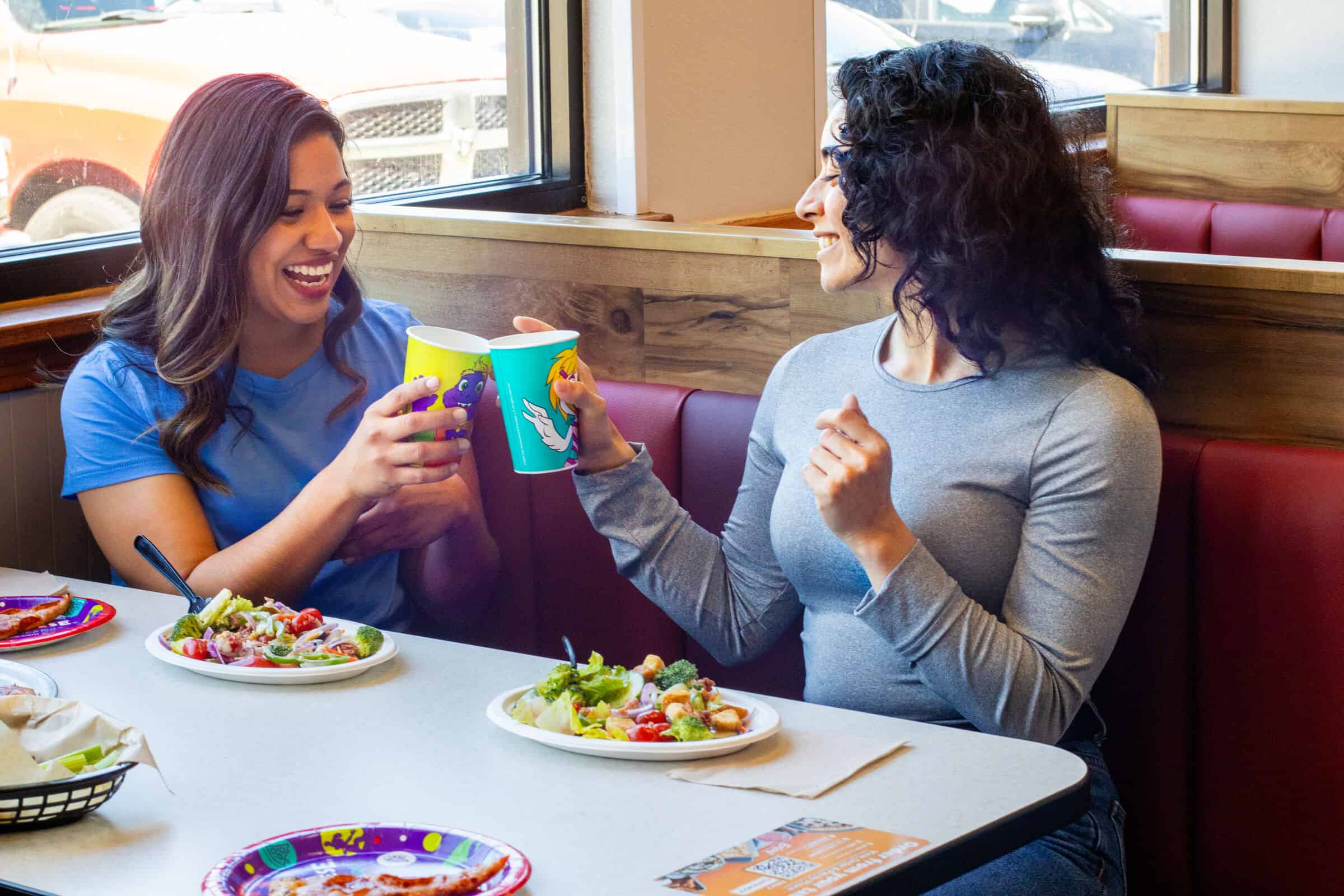 Two women at a Chuck E. Cheese table clink colorful cups by a window, salads and pizza on the table during a kids birthday party.
