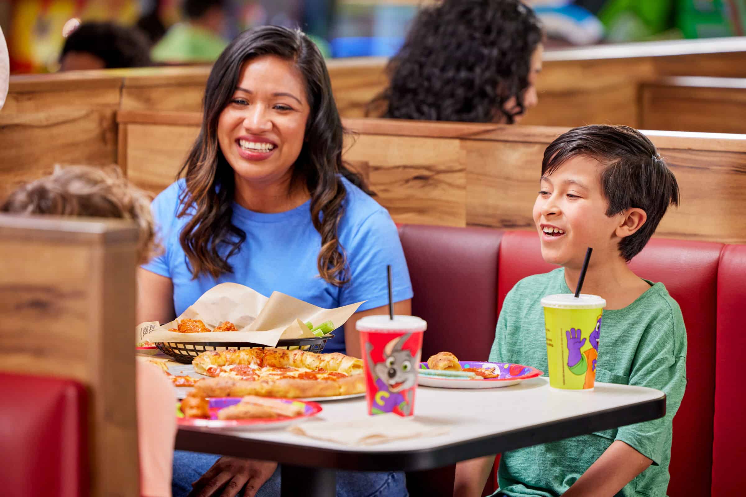 A woman and young boy smile at a Chuck E. Cheese booth sharing pizza and drinks, Fun Pass members.