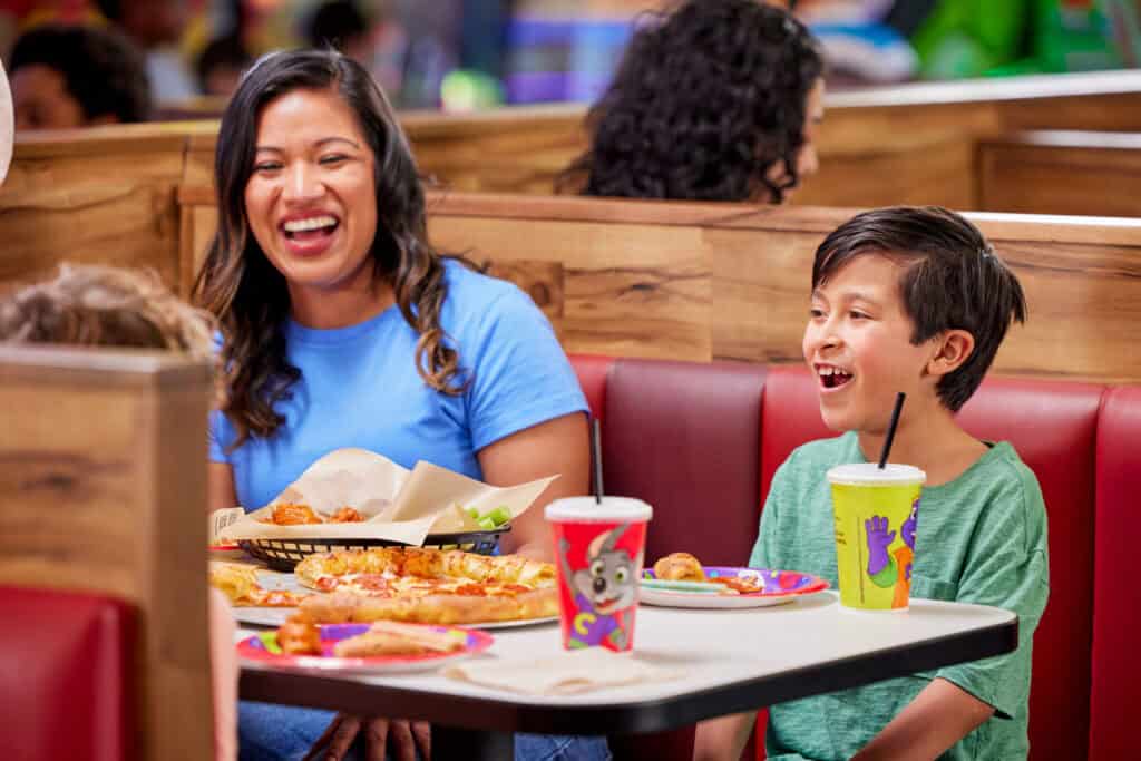 A woman and young boy enjoy pizza at a Chuck E. Cheese booth, smiling during a kids birthday party; other families in background.