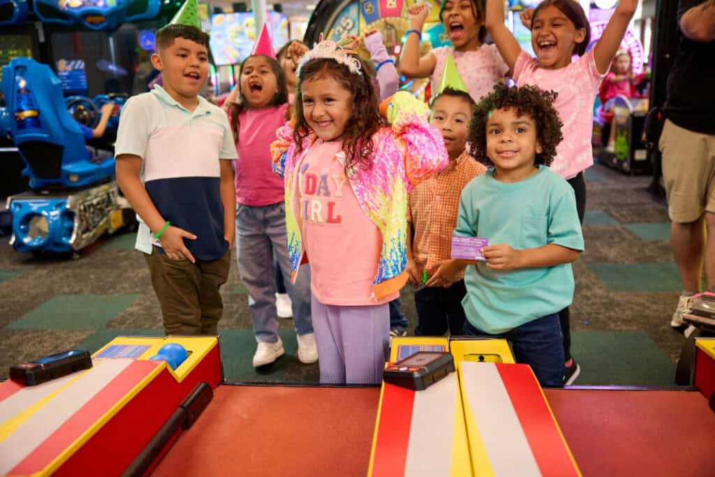 Birthday girl wearing a crown at Chuck E. Cheese, friends cheering around her near the skee-ball games, pure joy on her face