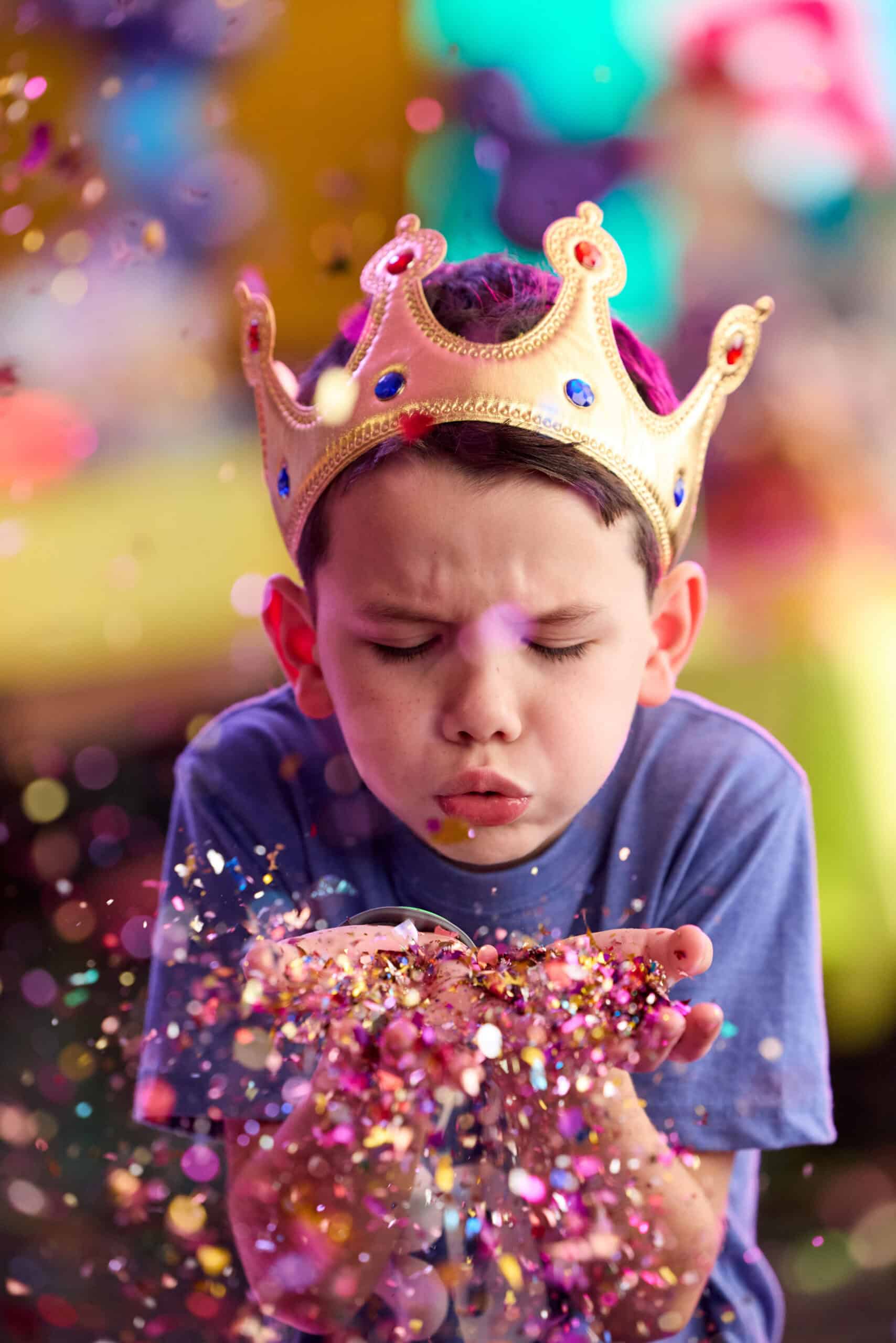 A boy in a gold crown blows confetti at a Chuck&nbsp;E.&nbsp;Cheese birthday party, with festive lights and arcade games in the background.