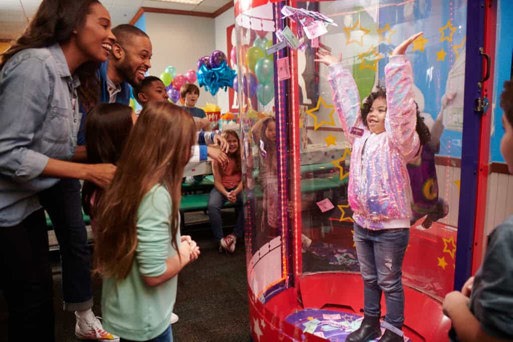 Birthday child inside the Ticket Blaster at Chuck E. Cheese as excited kids and adults cheer from outside.