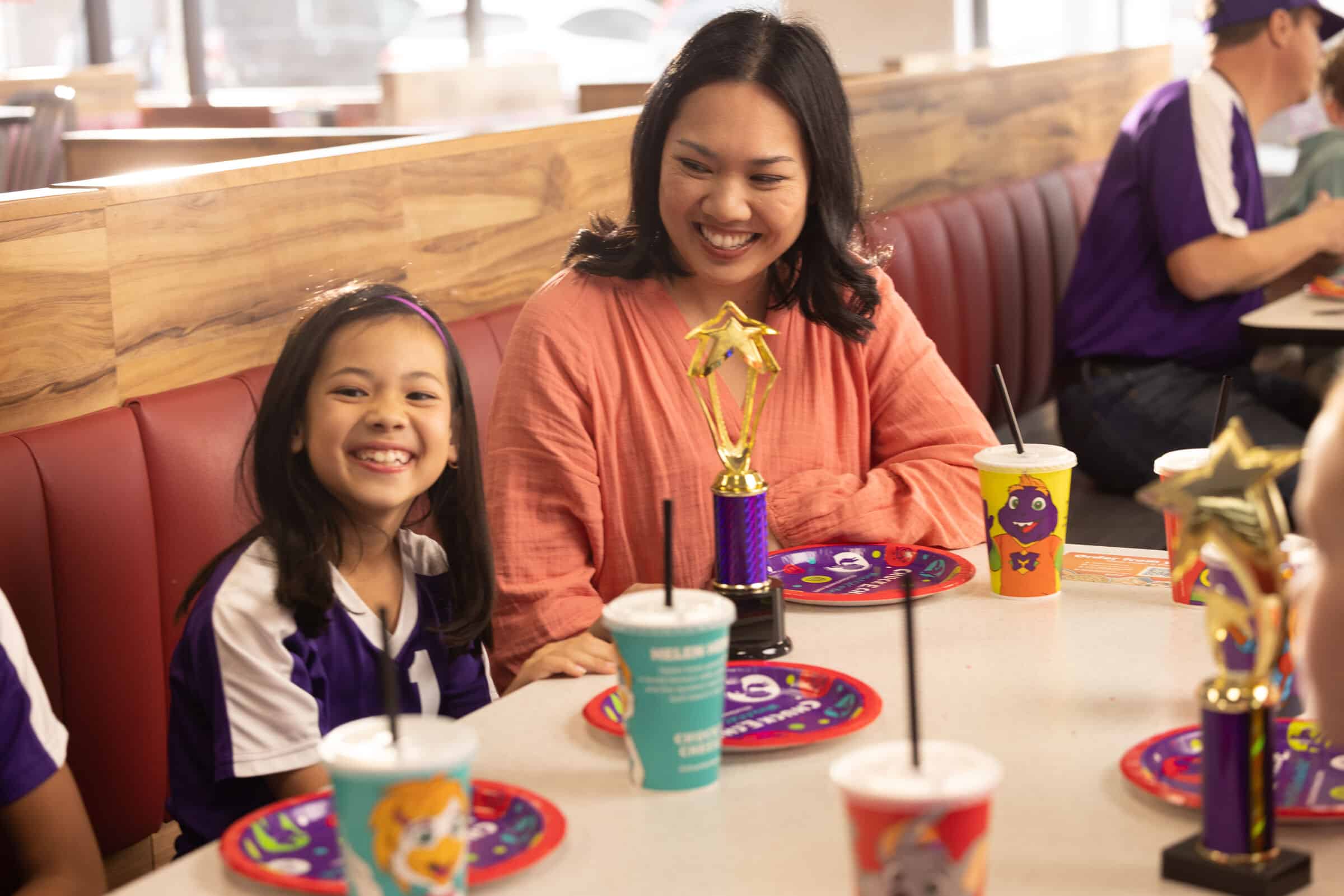 A woman and girl in sports uniforms celebrate at Chuck E. Cheese with pizza and colorful drinks