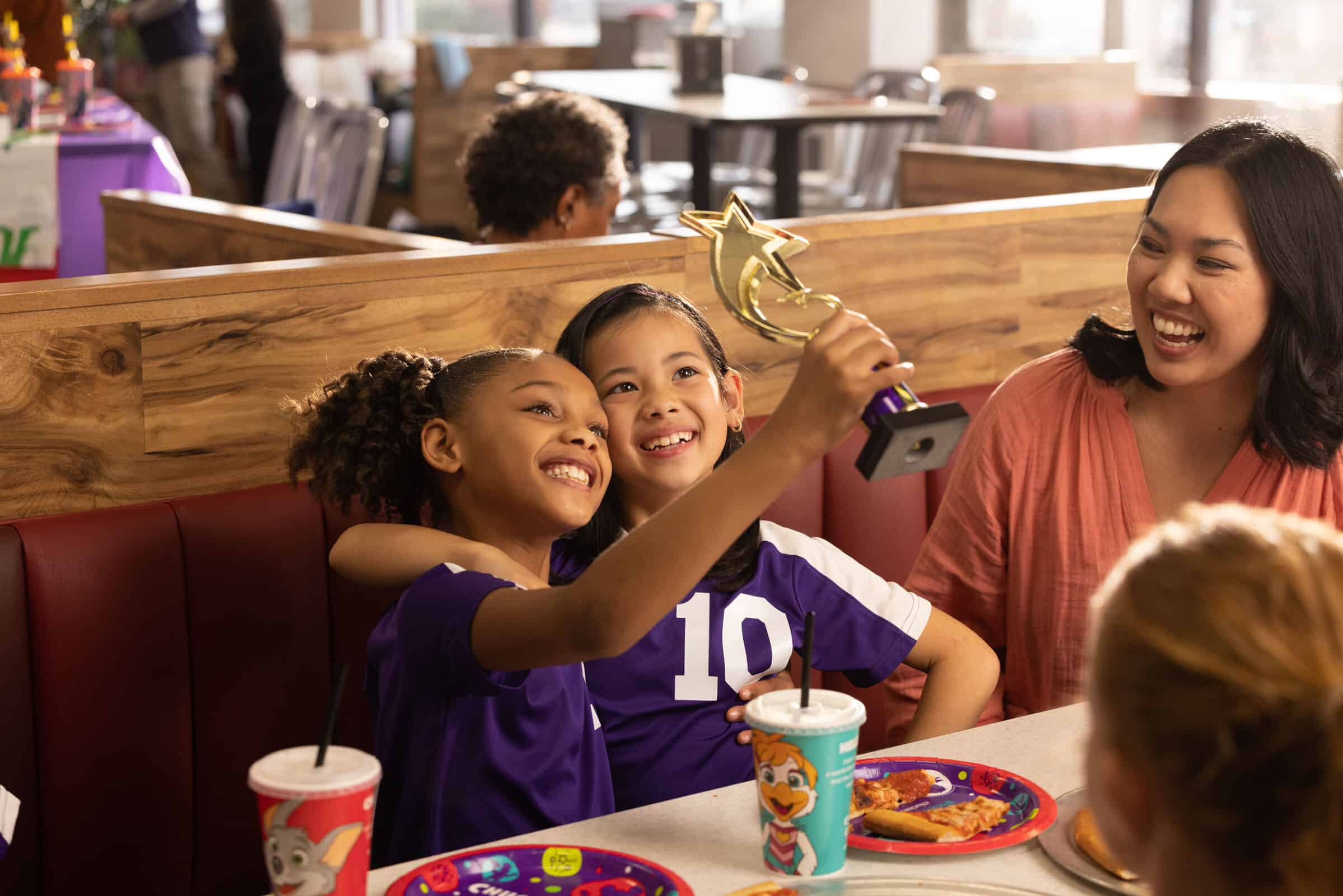 Two girls in purple soccer uniforms with a trophy and an adult at a Chuck E. Cheese team celebration