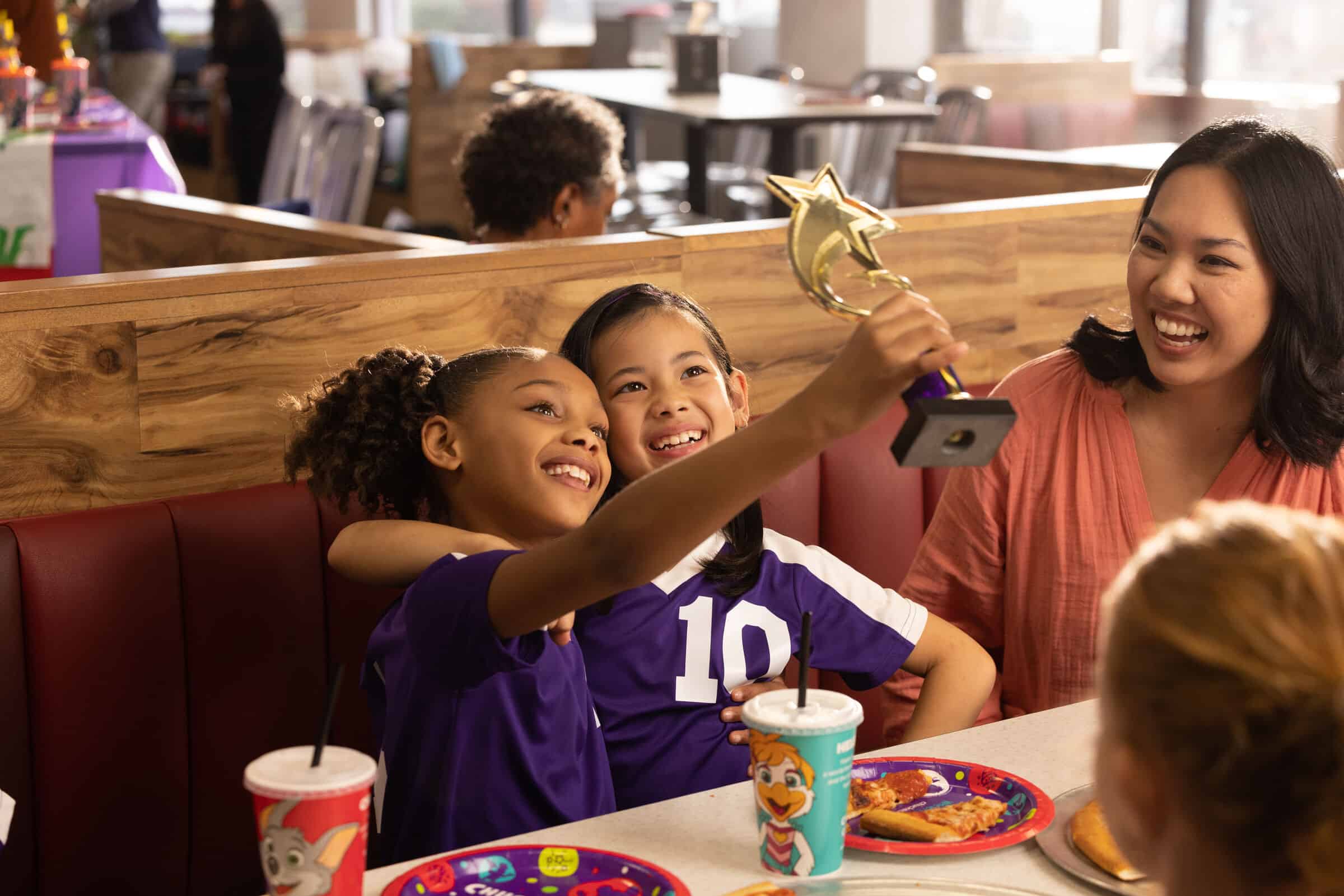 Two girls in purple soccer uniforms take a selfie with a trophy at Chuck E. Cheese — no gear required