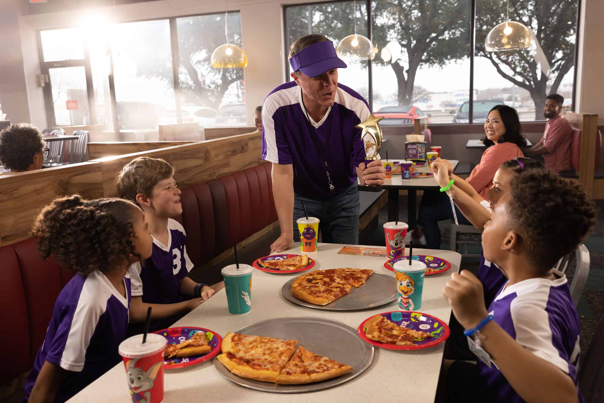 A youth soccer coach and four kids in purple jerseys celebrate with pizza at Chuck E. Cheese after their season