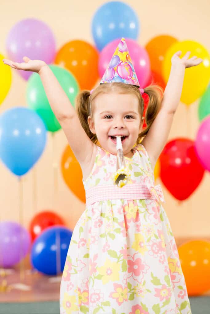 Young child, ages 3–5, in party room at Chuck&nbsp;E.&nbsp;Cheese birthday party with balloons, party hat, and a party blower.