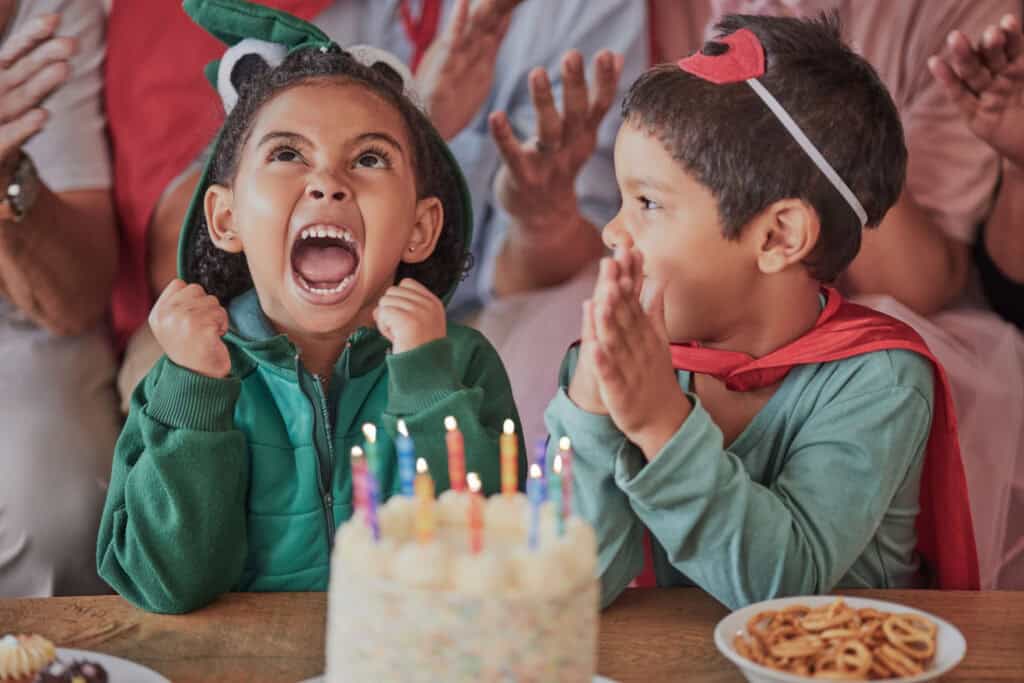 Birthday party in Chuck&nbsp;E.&nbsp;Cheese party room: 2 young kids in costumes, cake and snacks on table, family guests clapping around them.