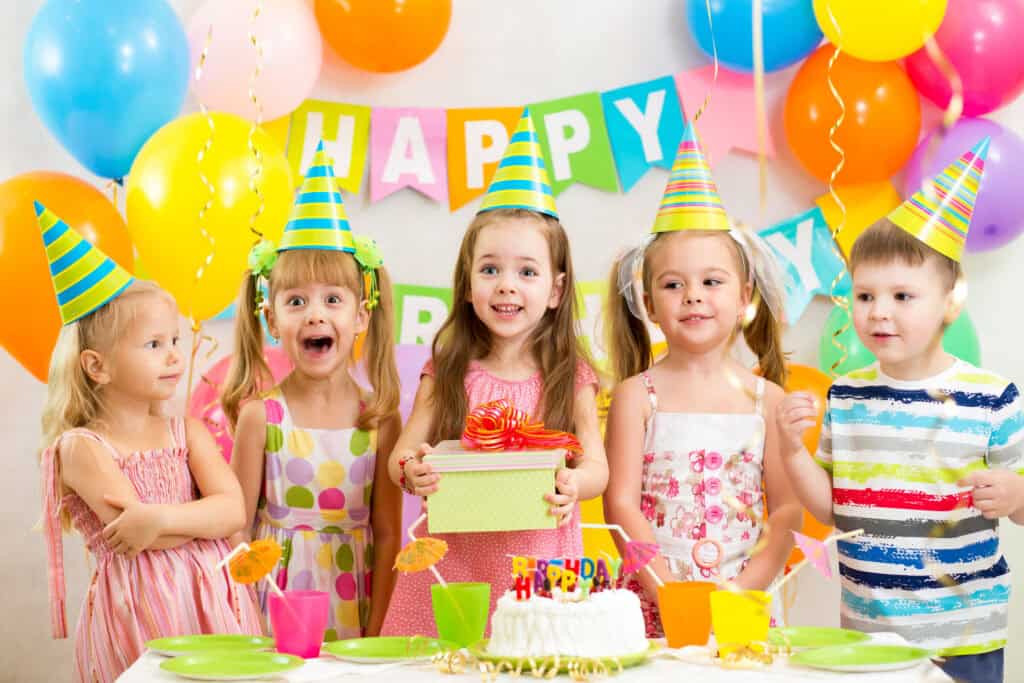 Kids ages 5–8 celebrate in Chuck&nbsp;E.&nbsp;Cheese party room with birthday cake, decorations, and arcade games in background.