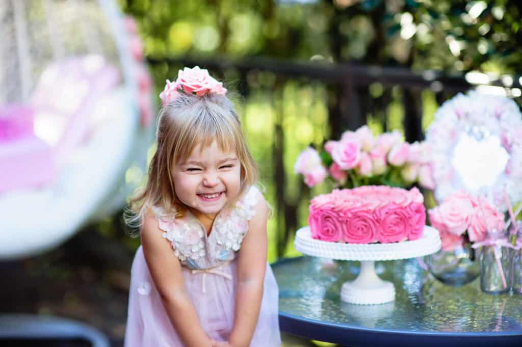 Birthday party at Chuck&nbsp;E.&nbsp;Cheese: girl (ages 4–6) in pink dress smiles by rose cake on glass table, party room with flowers.