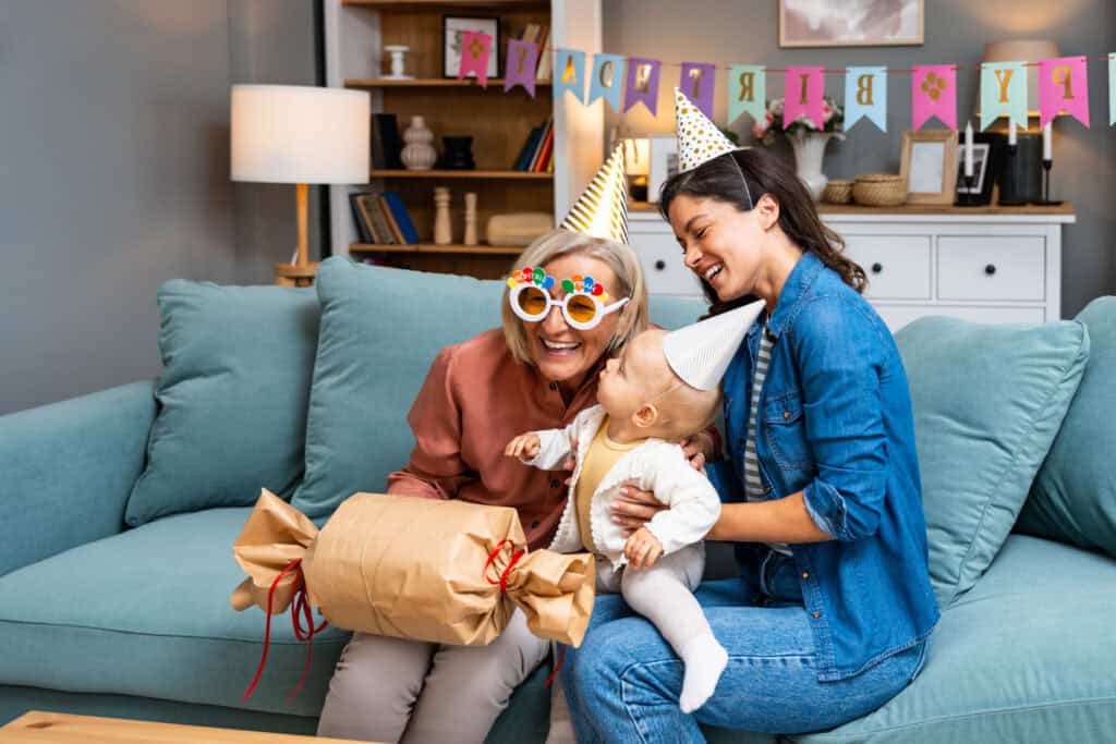 Older woman with fun glasses, baby, and adult in party hats at a Chuck&nbsp;E.&nbsp;Cheese birthday with gift and decorations nearby.