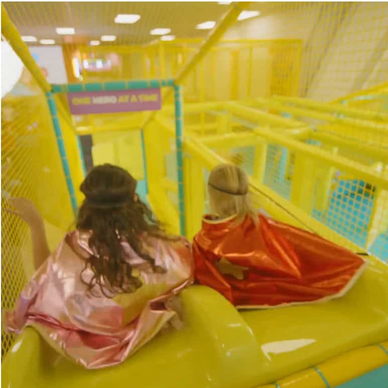 Two kids in capes and headbands sit atop a yellow indoor slide inside the Chuck E. Cheese Superhero Playground.