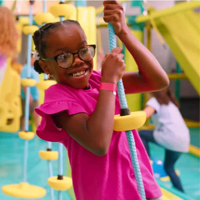 Young girl in glasses and pink shirt smiles holding a rope at Chuck E. Cheese, with kids playing in the background.