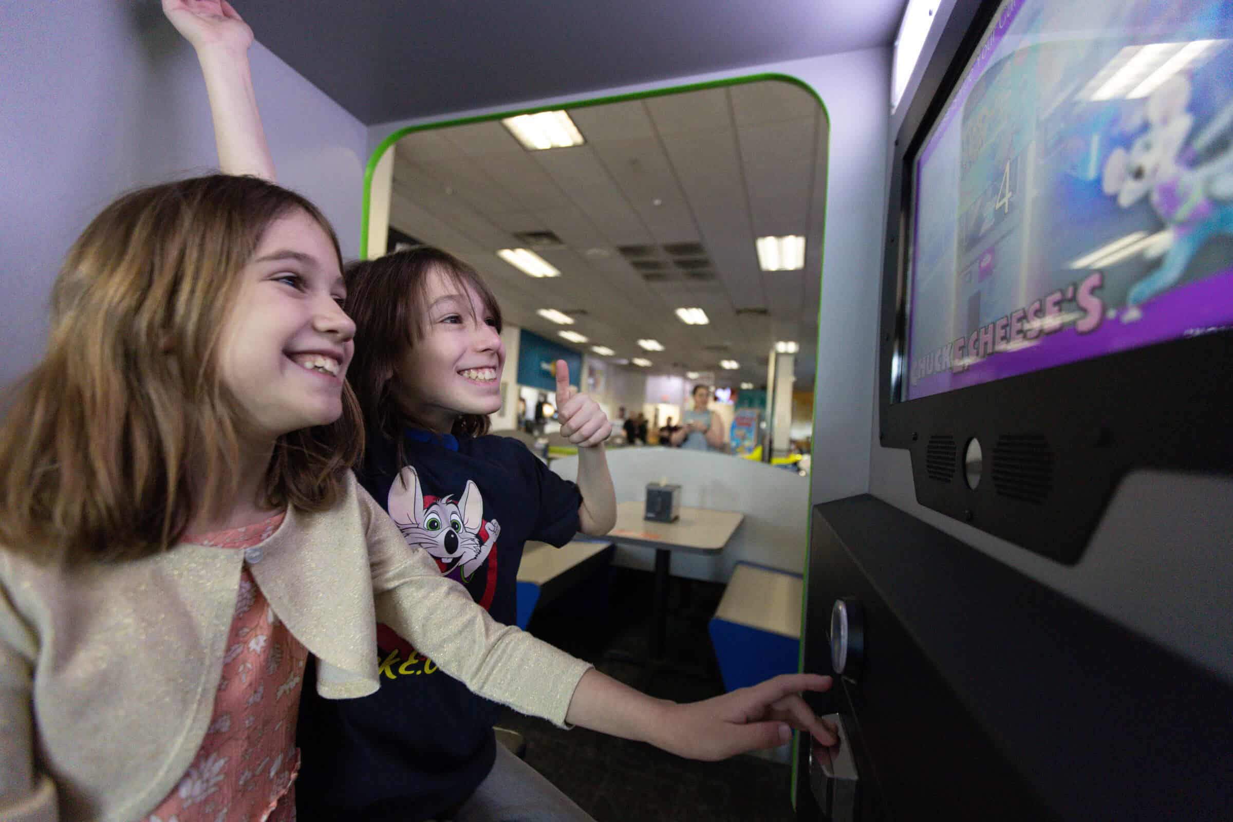 Two happy kids playing an arcade game at Chuck E. Cheese, one giving a thumbs up
