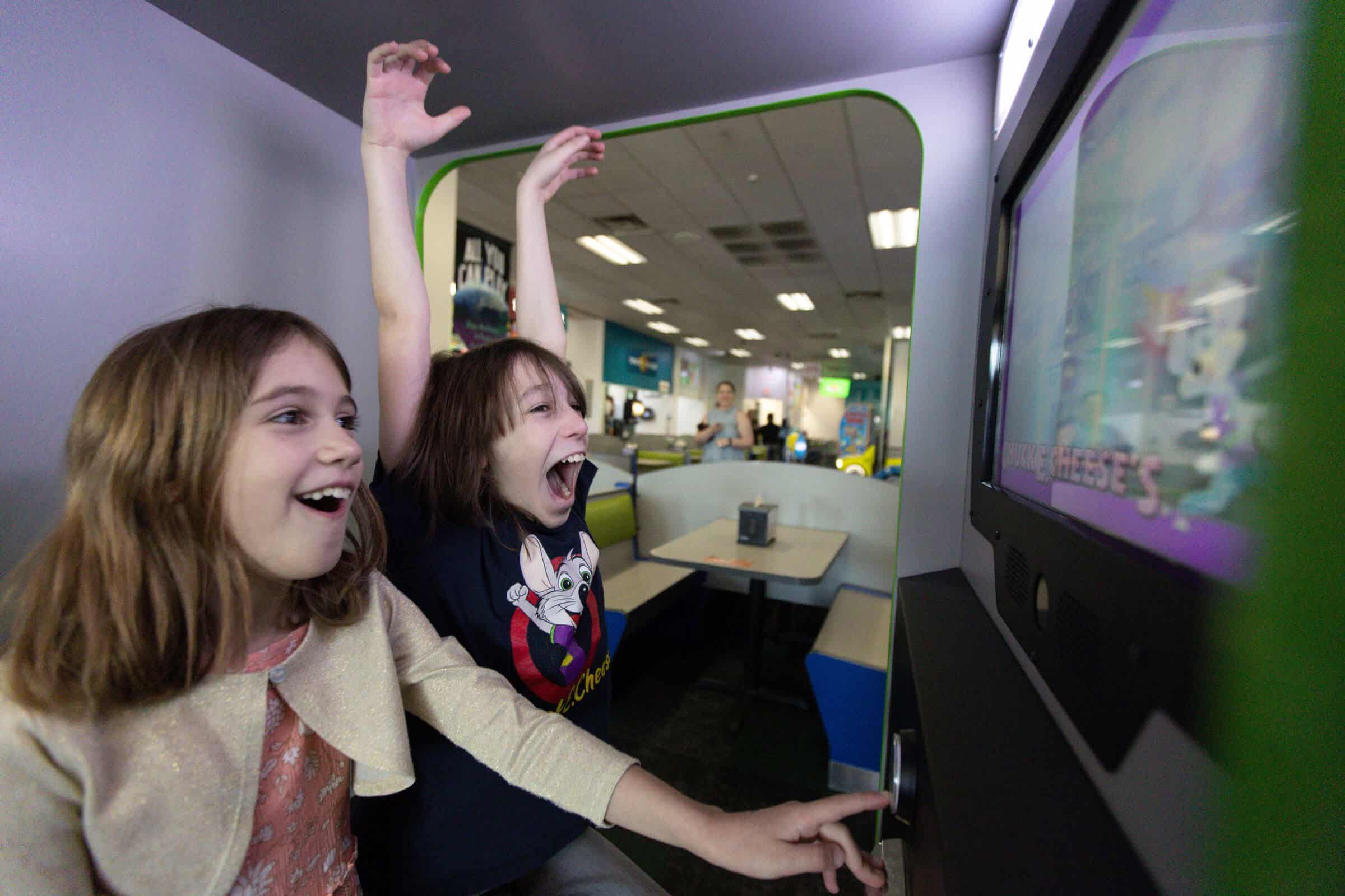 Two excited girls playing arcade games at Chuck E. Cheese, one cheering with arms raised