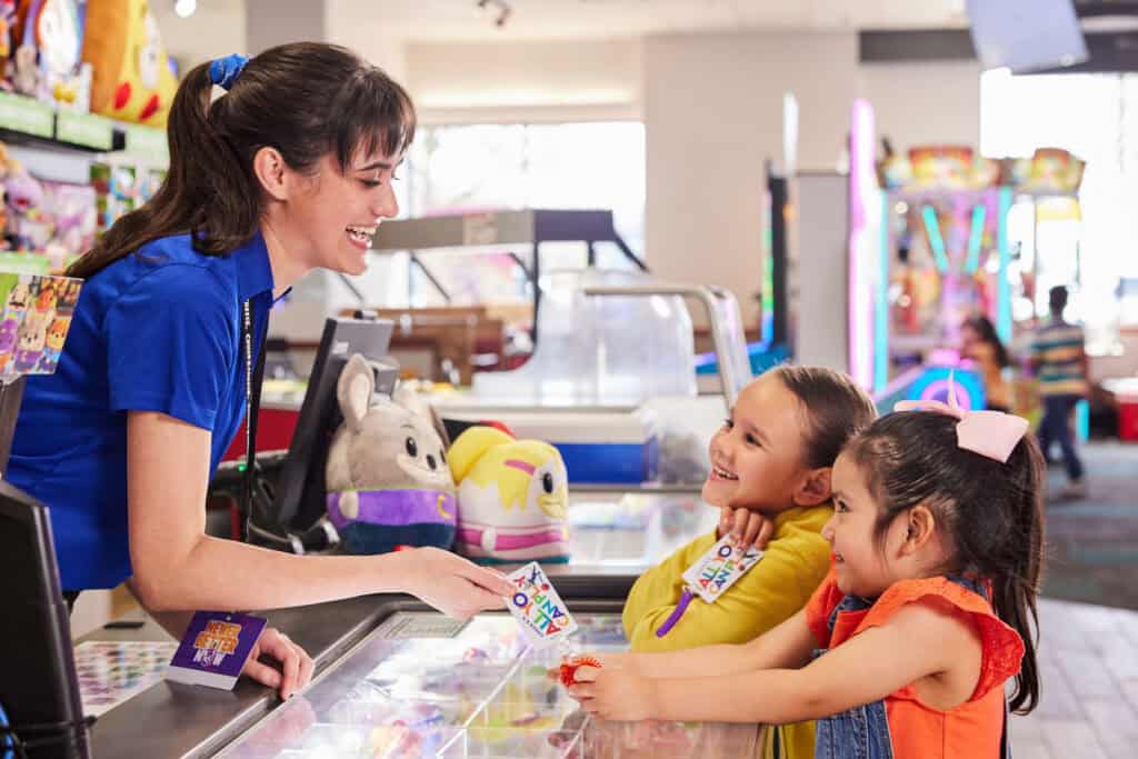 Chuck E. Cheese team member handing a Fun Pass card to two excited girls at the prize counter.