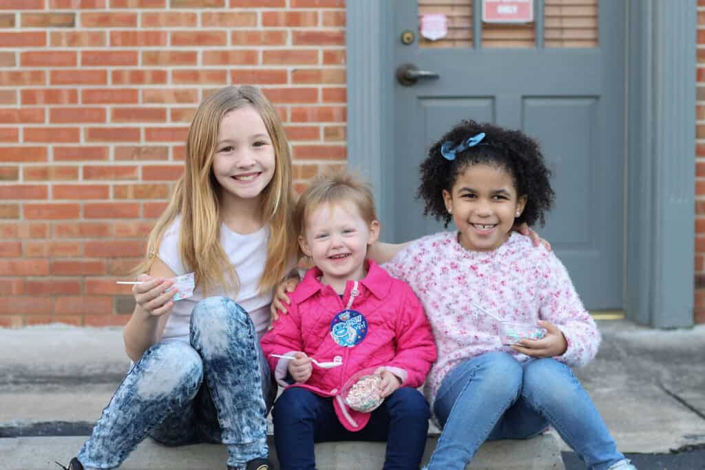 Three happy girls of different ages sitting together on steps outside Chuck E. Cheese, holding birthday decorations and smiling.