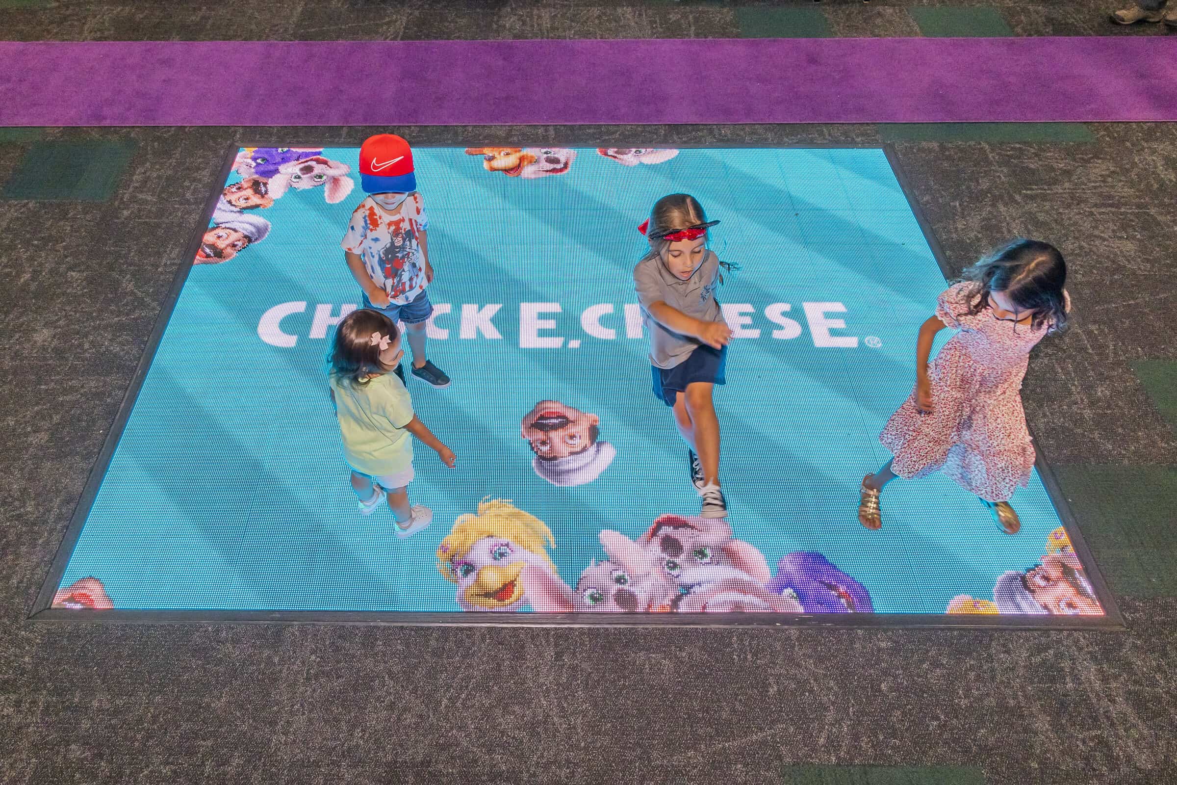 Kids playing on an interactive light-up dance floor with Chuck E. Cheese characters.