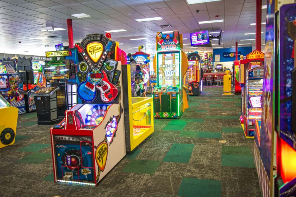 Brightly lit arcade floor at Chuck E. Cheese with various colorful games and dining area in the background.
