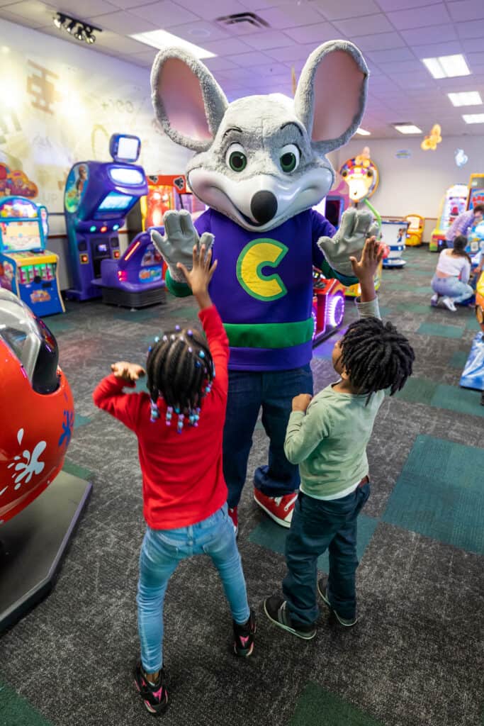 Chuck E. Cheese giving high-fives to two excited children in the arcade.