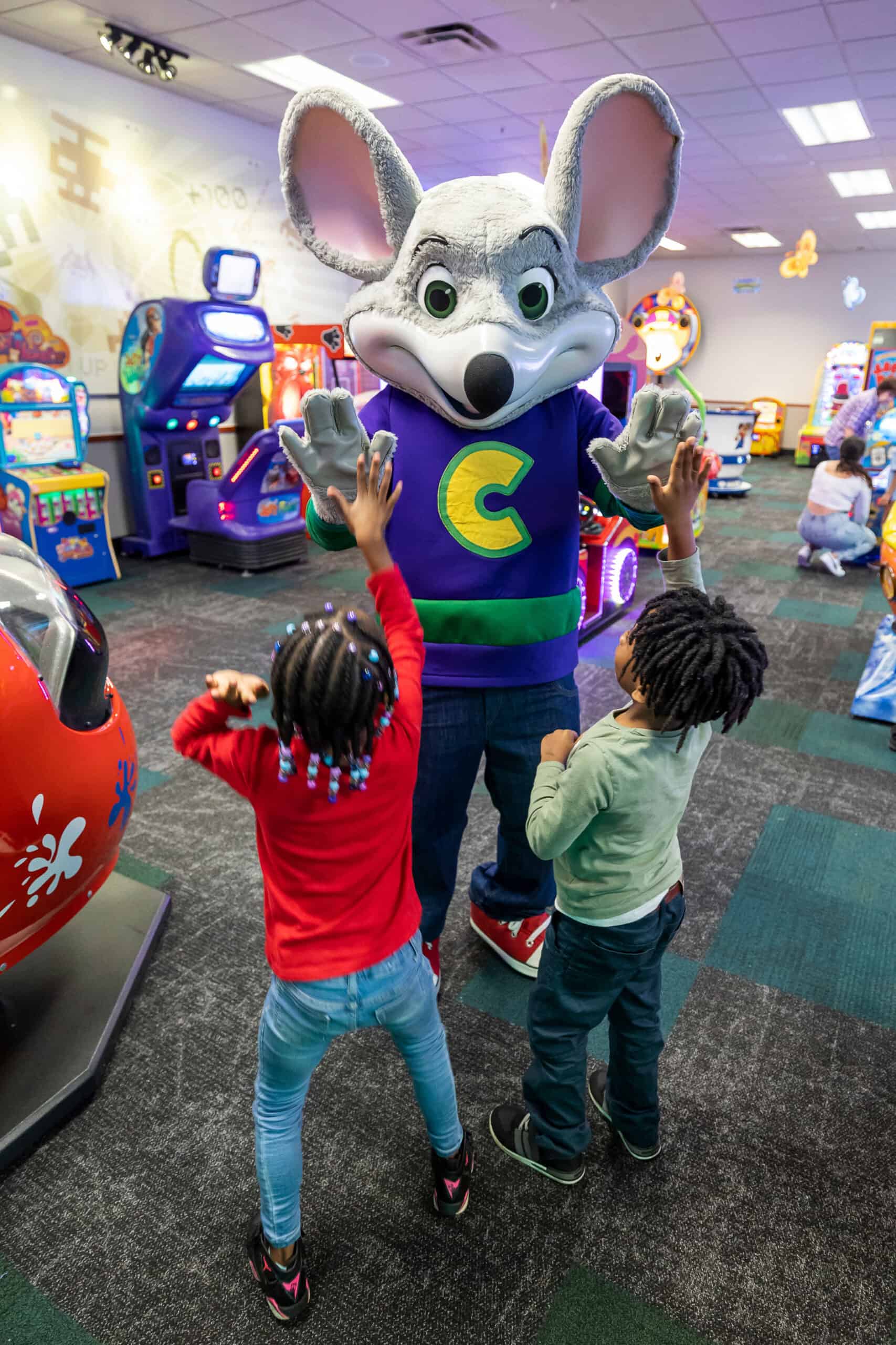 Chuck&nbsp;E.&nbsp;Cheese giving high-fives to two excited children in the arcade.