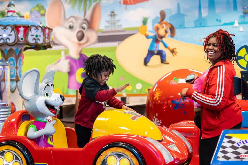 Child playing in a Chuck E. Cheese ride-on game while a woman smiles nearby.