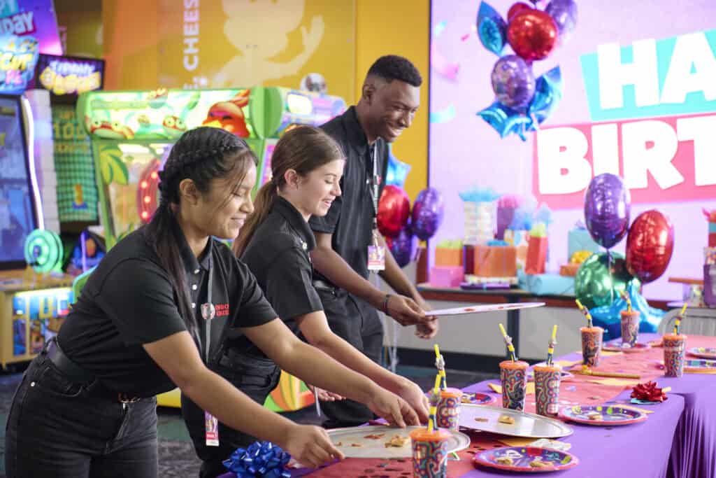 Chuck E. Cheese team members setting up a colorful birthday party table with pizza and drinks.