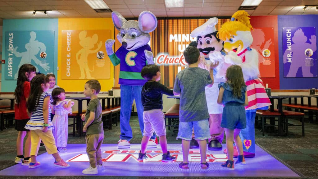 Chuck E. Cheese, Pasqually, and Helen Henny greeting a group of excited children on a light-up dance floor.