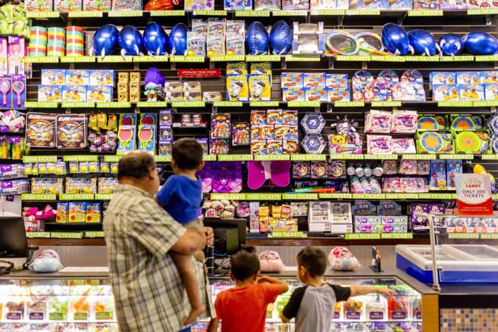 Father holding a young child while two other children look at prizes at the Chuck E. Cheese prize counter.