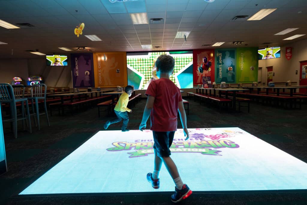 Kids playing on the interactive dance floor at Chuck E. Cheese, with character banners in the background.