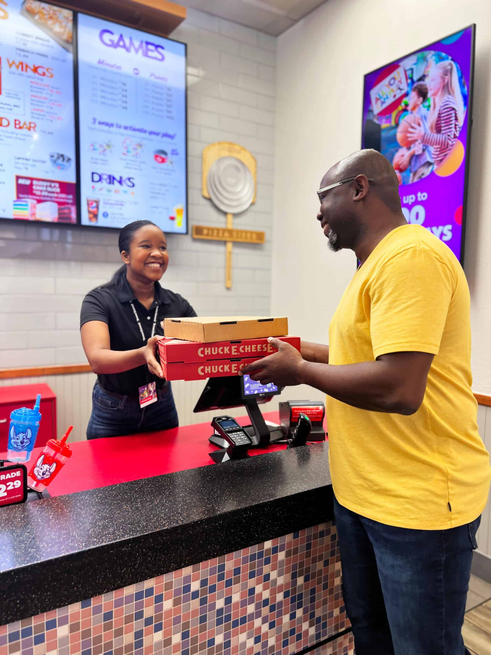Chuck E. Cheese team member handing two pizzas to a smiling adult customer at the counter