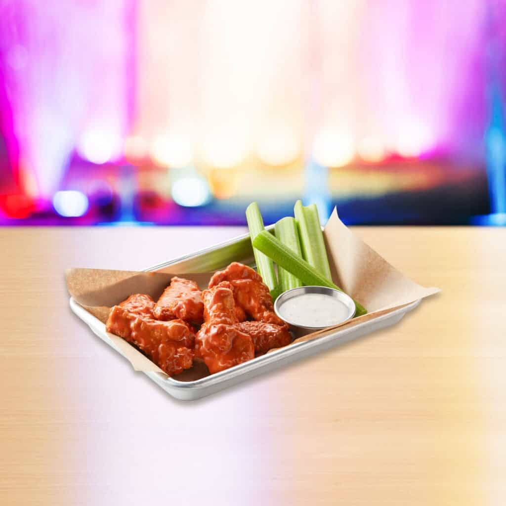 Buffalo chicken wings with celery and ranch on a party room table at Chuck E. Cheese family fun center, arcade lights behind.