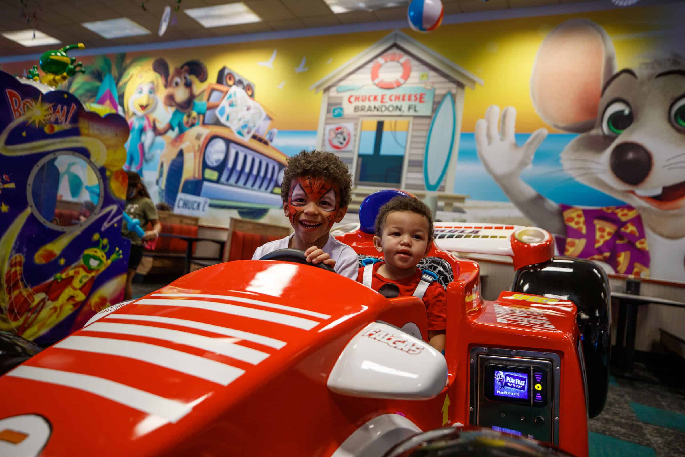 Two young boys smiling in a red race car arcade game at Chuck E. Cheese.