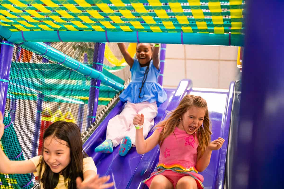 Three girls laughing and sliding down a purple slide in the Chuck E. Cheese play structure.